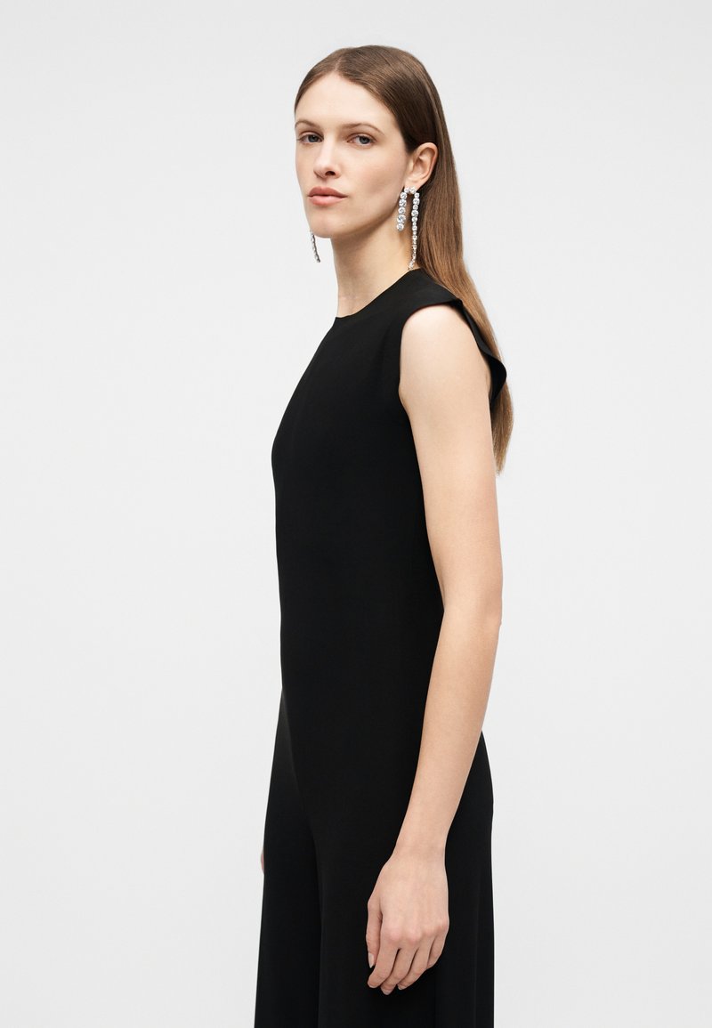 Femme aux longs cheveux bruns et lisses, portant une robe noire sans manches et de longues boucles d'oreilles argentées pendantes, se tenant devant un fond clair uni.