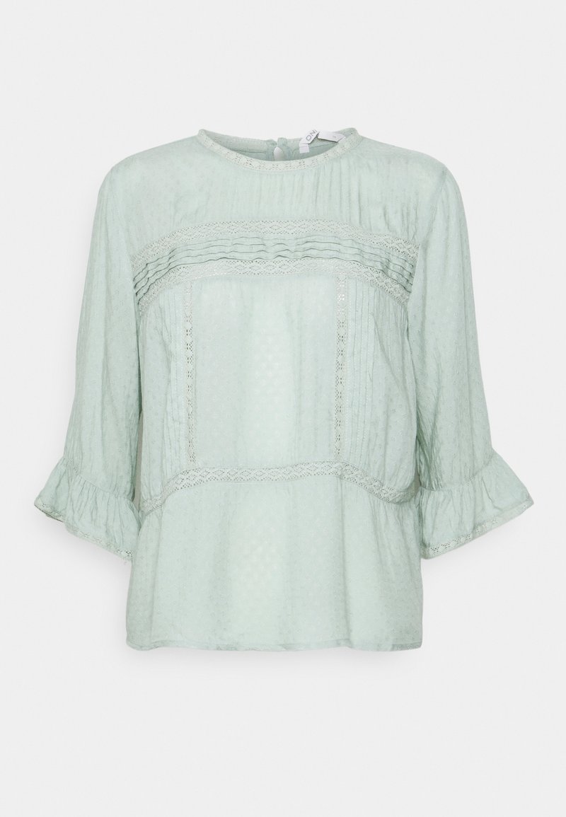 Jasnozielona bluzka z rękawami 3/4 i rozszerzanym kształtem, z fakturowanej tkaniny, pionowymi fałdami, koronkowymi dodatkami oraz okrągłym dekoltem z zapięciem na guzik z tyłu.