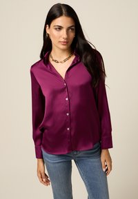 Paarse satijnen blouse met een gevelde kraag, knoopsluiting, lange mouwen en een relaxte pasvorm, gecombineerd met blauwe jeans. Accenten van een gouden ketting.