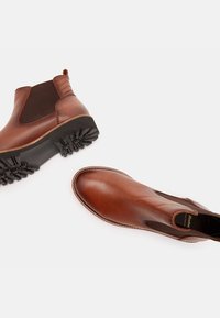 Ruskeat nahkaiset Chelsea-saappaat, joissa on joustavat sivupaneelit ja teksturoitu musta kumipohja; sileä pinta ja kontrastitikkaus.