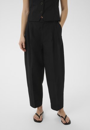 Femme portant un pantalon noir taille haute à jambes larges, un gilet noir sans manches boutonné et des sandales noires à lanières, debout sur un fond blanc.