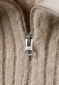 Beiger gestrickter Stoff mit strukturiertem Muster und sichtbaren Fasern, ausgestattet mit einem silbernen Reißverschluss-Zipper, der mit "WEEKDAY" markiert ist, neben den Zipperzähnen.
