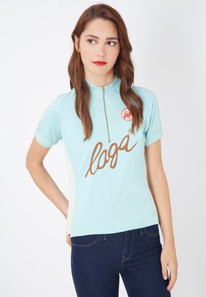 Top de corto azul claro con cuello con cremallera, que presenta un gráfico del logo marrón en la parte frontal y paneles laterales blancos. Textura de tela suave.