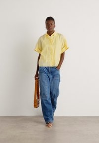 Camicia gialla a maniche corte con bottoni, taglio ampio, abbinata a jeans a gamba larga blu chiaro. Borsa marrone con tracolla tenuta nella mano sinistra.
