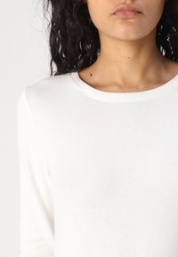 Top in cotone bianco a maniche lunghe con texture a coste, colletto rotondo e dettagli di cucitura sottili.
