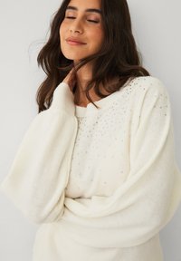 Cremfarbener Pullover mit lockerer Passform, der eine strukturierte Oberfläche und verstreute silberne Verzierungen am Ausschnitt und an den Schultern aufweist.