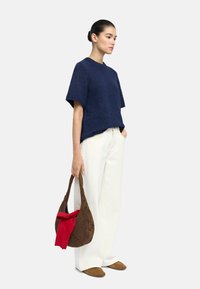Niebieska, teksturowana bluzka z krótkim rękawem, beżowe spodnie o szerokich nogawkach, brązowa pleciona torba z czerwonym akcentem. Model stoi na neutralnym tle.