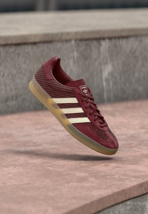Bordowy sneaker Adidas z beżowymi paskami i gumową podeszwą unoszący się lekko nad różową kamienną powierzchnią i szarym kamiennym tłem.