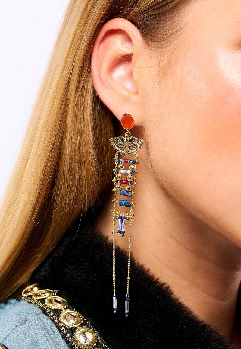 Boucles d'oreilles en ton doré avec une pierre orange et plusieurs fils suspendus de perles colorées, incluant des détails bleus, rouges et blancs.