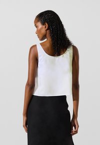 Ženska s kodrastimi lasmi, oblečena v belo neformalno zgornjo majico brez rokavov in črno krilo, obrnjena stran proti preprosti svetli podlagi.