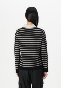 Kvinna med långt mörkt hår vänd bort, iförd en svart och vit randig långärmad tröja och svarta cargobyxor.