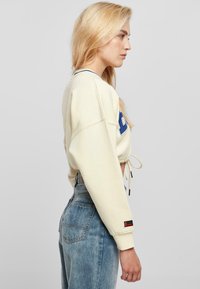 Light beige cropped sweatshirt with blue logo detailing, drawstring at the hem, and oversized sleeves paired with high-waisted blue jeans.