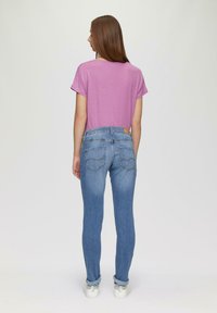Camisa de manga corta color púrpura claro combinada con jeans de mezclilla azul ajustados que cuentan con bolsillos traseros y un sutil diseño de costura. Zapatillas blancas visibles.