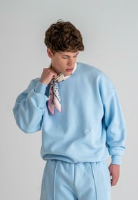 Hellblaues Sweatshirt mit lässigem Schnitt, gerippten Bündchen und gesticktem Logo. Kombiniert mit einem gemusterten Schal in pastelligen Farben.