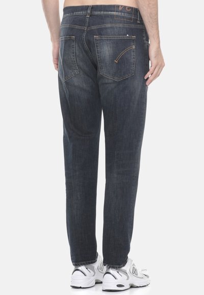 Uomo che indossa jeans blu scuri e sneakers bianche, visto da dietro su uno sfondo chiaro.