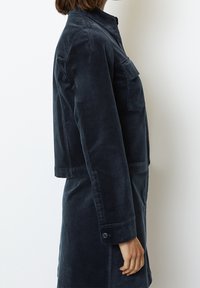 Tmavě modrá manšestrová bunda střední délky s límcem, bočními kapsami a knoflíkovými rukávy. Má volný střih a hladkou texturu.