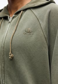 Κοντινό πλάνο ενός ατόμου που φοράει ελιά πράσινο φούτερ με φερμουάρ και κορδόνια, με το λογότυπο Champion κεντημένο στο στήθος.