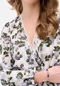 Blumenbluse in Weiß, Grün und Lila mit einem Spitzenbesatz am Ausschnitt und gerafften Ärmeln. Das Armband besteht aus blauen, türkisfarbenen, beigen und lila Perlen.