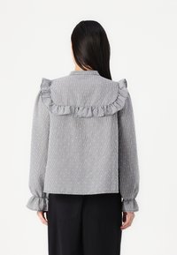 Grå rutigt blus med volangkrage och manschetter, med en texturerad tyg och subtila prickar. Långa ärmar och avslappnad passform.