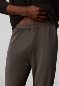 Homme portant un pantalon de détente gris Calvin Klein remontant un t-shirt noir pour révéler le logo de la ceinture.