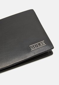 Portefeuille en cuir noir avec une surface texturée, orné d'un logo "GUESS" en argent et d'un design minimaliste. Compact avec une forme élégante.