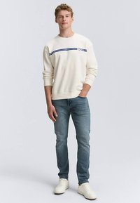 Krémově zbarvená mikina s tmavě modrým proužkem, v kombinaci se světle modrými džínami a bílými teniskami, s volným střihem a žebrovanými manžetami.