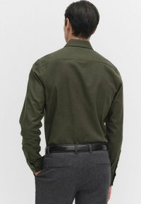 Oliva vihreä pitkähihaisuus paita klassisella kauluksella, näkyy takaa, yhdistettynä harmaisiin muotoleikkaus housuihin ja mustaan vyöhön.