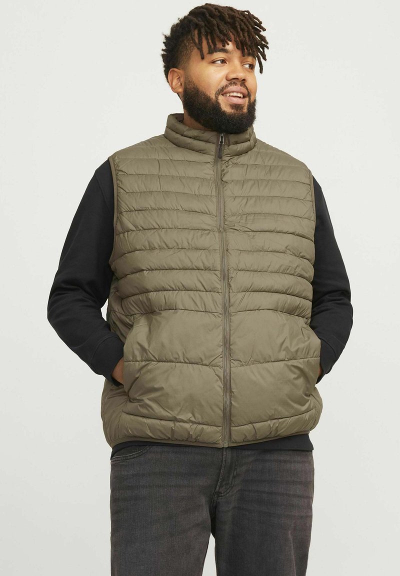 Gilet rembourré en vert olive, avec un col montant, une fermeture éclair à l'avant et des poches latérales. Porté par-dessus une chemise à manches longues noire.