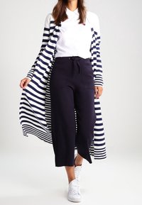 Dlouhý pruhovaný kardigan v námořnické modři a bílé, přes bílou košili, spárován s námořnickými culottes a bílými teniskami. Hladké látky, ležérní design.