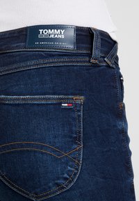 Mörkblå denimjeans med en synlig bakficka med kontrastsömmar, en läderetikett med "TOMMY JEANS" och en liten logotypdetalj.
