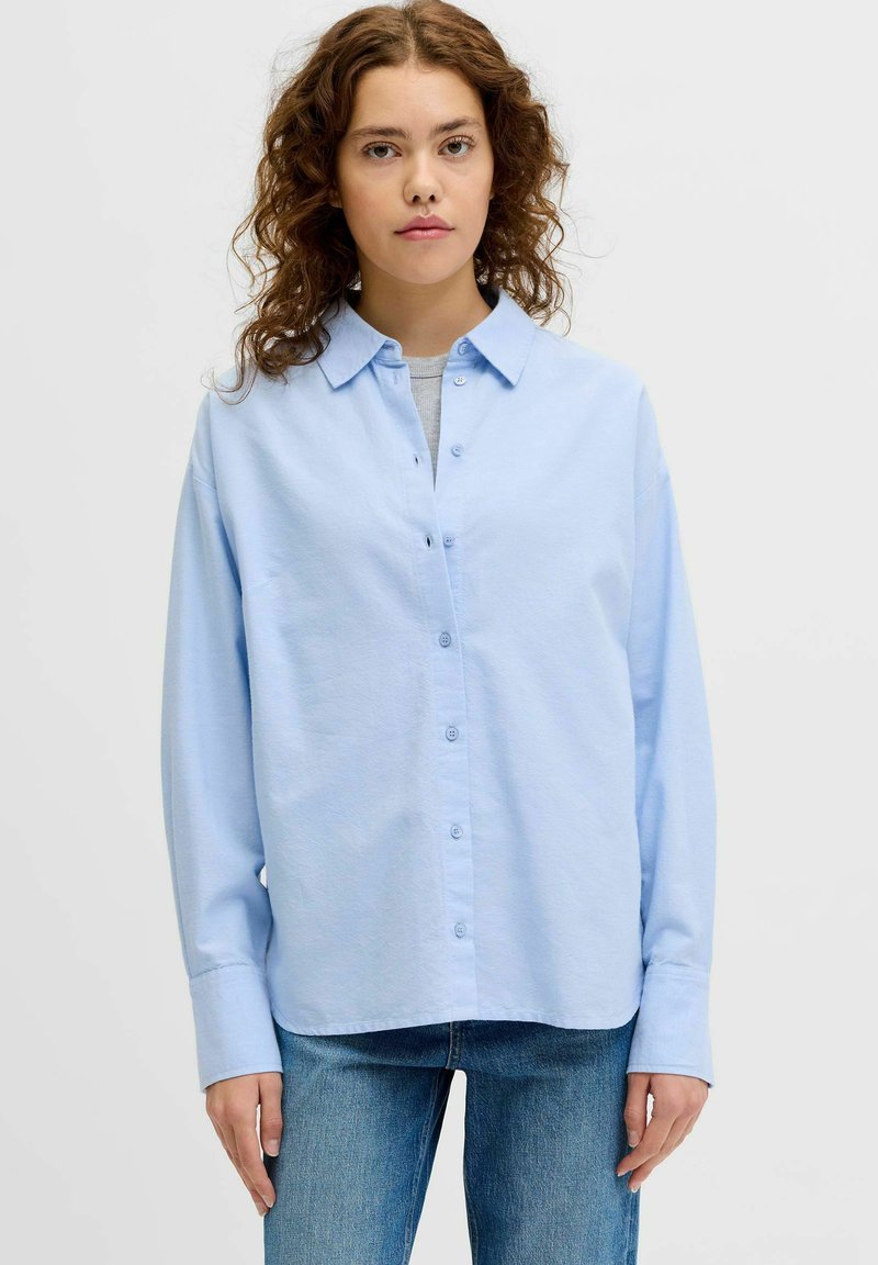 Jonge vrouw met krullend bruin haar die een lichtblauw overhemd draagt over een grijs shirt en blauwe spijkerbroek, staand tegen een effen achtergrond.