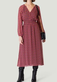 Robe longue rose avec un motif géométrique, décolleté en V et taille élastique. Manches longues transparentes associées à des bottes noires arrivant au genou et un petit sac noir.