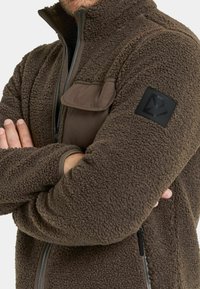 Ruskea fleece-takki, jossa on korkea kaulus ja vetoketju. Siinä on rintatasku ja musta logo-patch hihassa. Kankaan rakenne on pehmeä ja mukava.