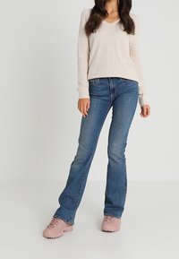Pull beige clair à manches longues avec col en V, jean bleu évasé et chaussures lacées rose pâle. La texture semble douce ; les matériaux sont décontractés.
