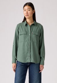 Groen blouse met knopen, voorzien van twee borstzakken, lange mouwen en een kraag. Zachte stof, iets losse pasvorm, met zilveren knopen en manchet.