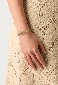 Bracciale d'oro con design a nodo intrecciato, indossato su una mano con un vestito di pizzo delicato e intricato, in beige, che mostra motivi di farfalle.