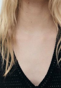 Schwarzes, gestricktes Oberteil mit V-Ausschnitt und einer strukturierten Musterung. Hellblondes Haar fällt über die Schultern und verdeckt teilweise den Ausschnitt.