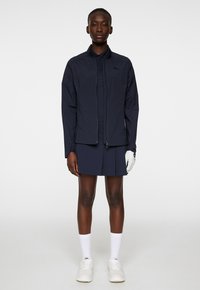 Marinblå vattentät jacka med dragkedja fram, långa ärmar och elastiska ärmslut, bärs över en marinblå polo-klänning. Vita strumpor och skor synliga.