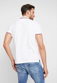 Polo bianco con accenti a righe rosse e blu navy nel colletto, maniche corte e vestibilità rilassata, abbinato a jeans di denim blu chiaro.