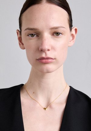 Colar com pendente em forma de coração dourado, numa corrente fina, usado contra um pescoço nu, com uma textura suave e design minimalista.