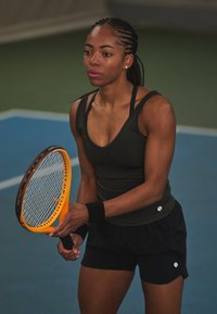 Idrettsutøver i en mørk tanktopp og svarte shorts, som holder en oransje tennisracket, med sterke armemuskler, på en blå bane.
