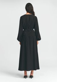 Schwarzes Maxi-Kleid mit Punkten, langen Ärmeln, geraffter Taille und fließendem Stoff, getragen mit schwarzen Absätzen. Rückansicht, glattes Material.