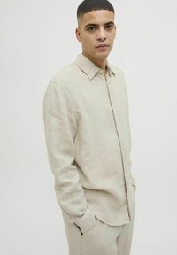 Camisa de lino beige claro con cuello abotonado, que presenta un corte relajado y mangas largas, llevada por dentro de pantalones de lino a juego.