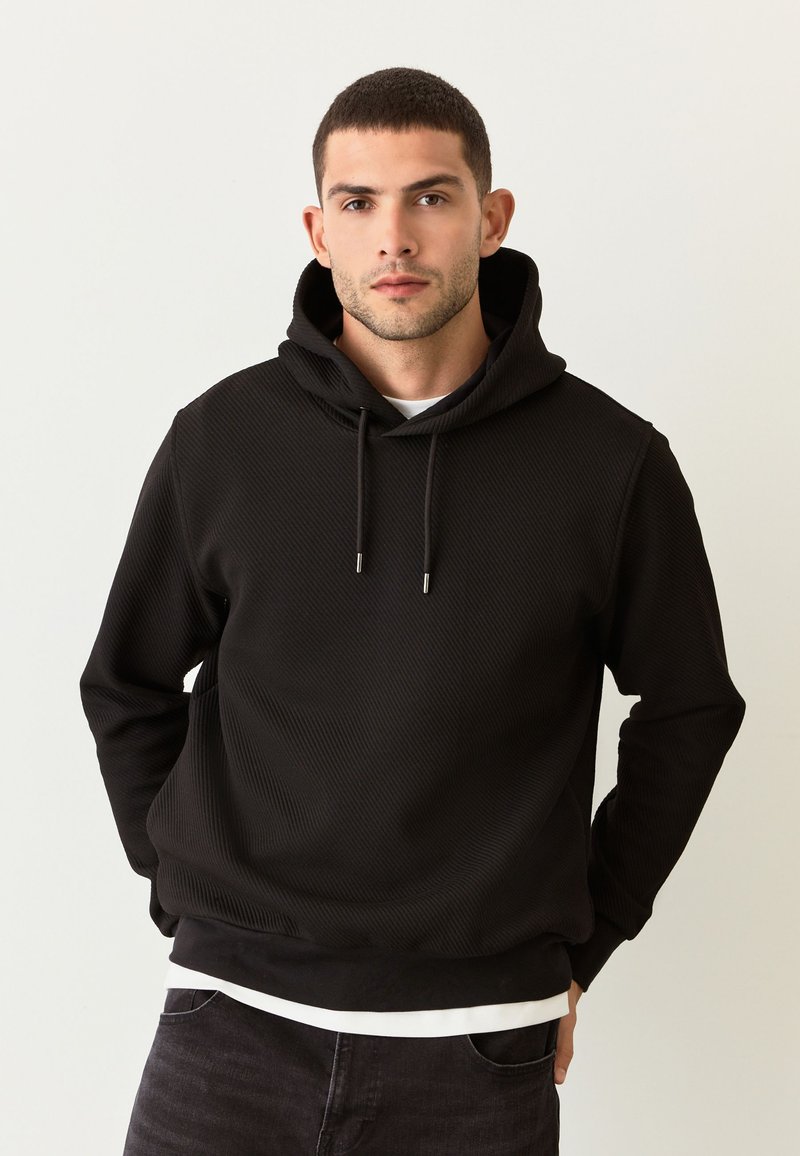 Felpa nera con cappuccio dal motivo strutturato, coulisse e orlo a coste sottile. Indossata sopra una camicia bianca, creando un contrasto con jeans scuri.