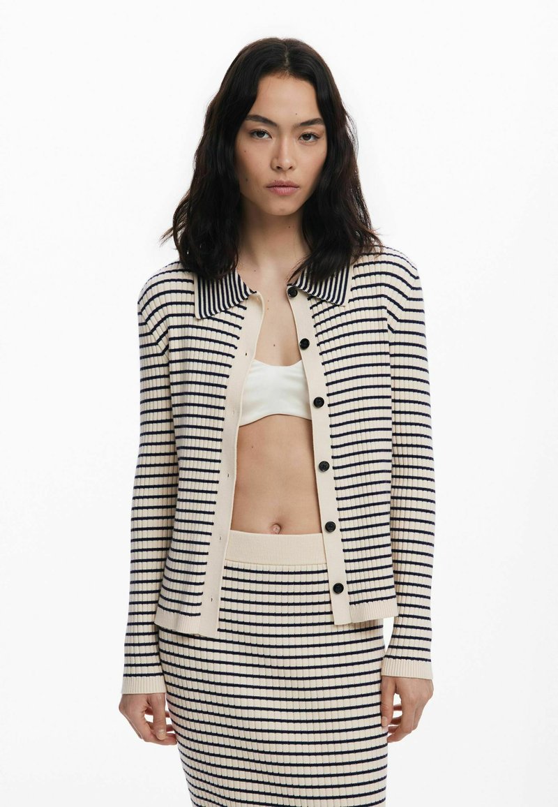 Cardigan crema con righe blu navy, texture a coste e scollatura a colletto. Presenta bottoni frontali e una gonna coordinata a righe.