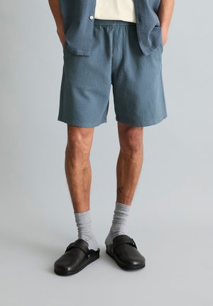 Man wearing blue textured shorts, matching open jacket, white shirt, gray socks, and black slip-on sandals, standing with hands in pockets.