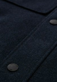 Caban de lână bleumarin cu un model herringbone, prins cu nasturi rotunzi negri, având o finisare a materialului netedă.