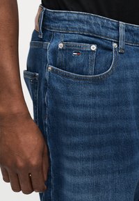 Κοντινό πλάνο σκούρου δέρματος χεριού δίπλα σε μπλε τζιν τζιν με ορατή μπροστινή τσέπη, μικρό λογότυπο σημαίας και καφέ δερμάτινο σημείο ζώνης.