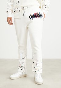 Hvite joggebukser med flerfarget malingssprut, elastisk linning med snøring, sideglidelåsdetailler, og brodert "Color Collective"-logo.