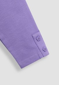 Violetti puuvillapala, jossa on sileä rakenne, ja jokaessa on kaksi kukkamuotoista nappia sekä ommeltu helmaresori.